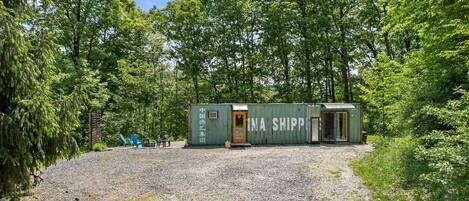 A small, green shipping container structure with a door, surrounded by gravel and trees.