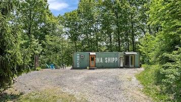 A small, green shipping container structure with a door, surrounded by gravel and trees.