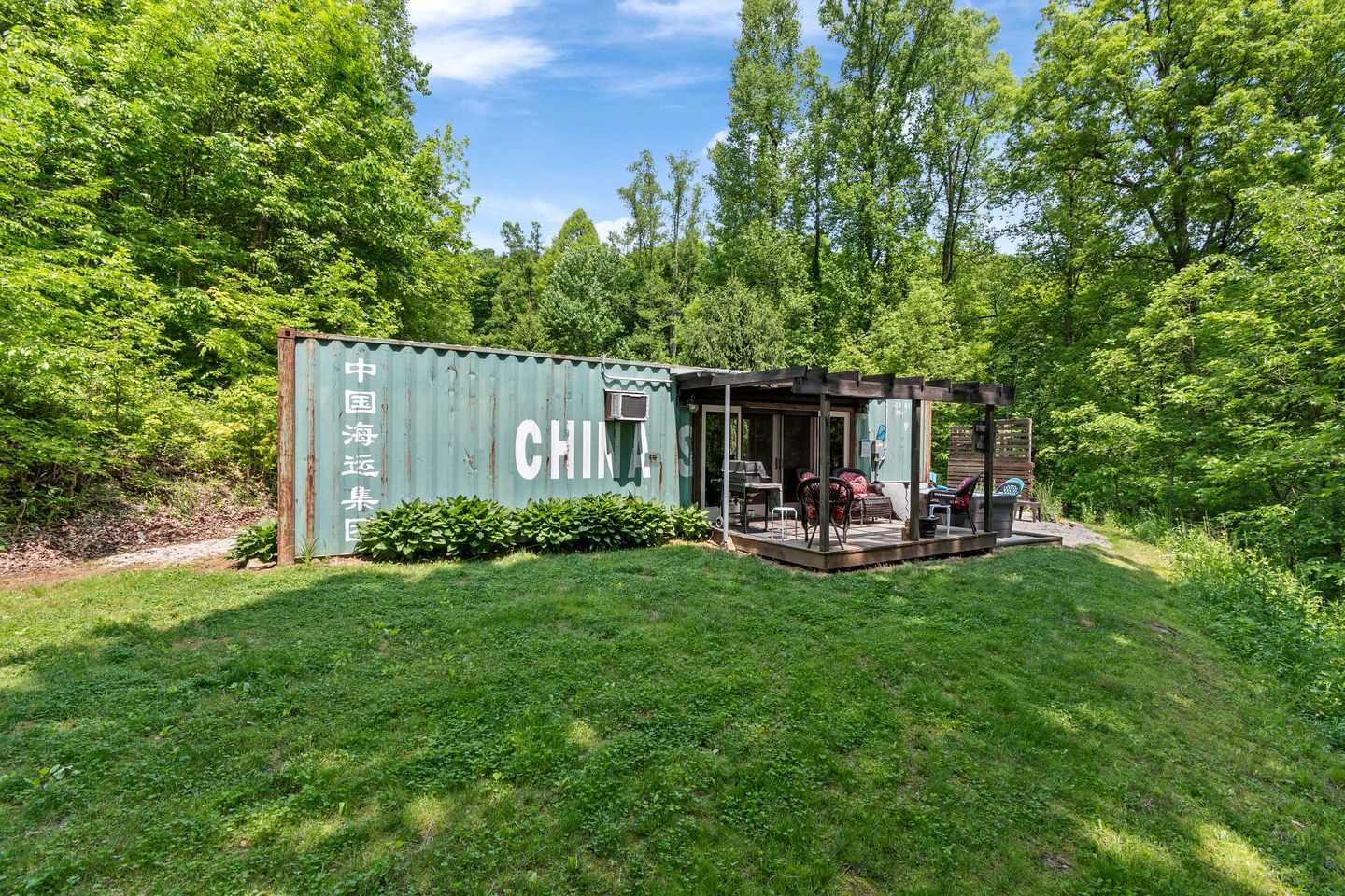 A green shipping container converted into a small outdoor structure with a covered patio, seating, and a grill area.