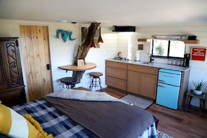 Una habitación compacta con una cama, una pequeña mesa de comedor con taburetes, una kitchenette con un refrigerador azul y una decoración rústica que incluye un tronco de árbol y un cráneo de oveja.