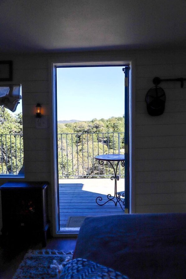 Una habitación con cama, una mesita, una silla, ventana y balcón con vistas a árboles y colinas.