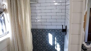 Un baño con paredes de azulejos blancos, zona de ducha, baldosas estampadas en el suelo y una ventana con cortina.