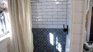 A bathroom with white tiled walls, a shower area, patterned tiles on the floor, and a window with a curtain.