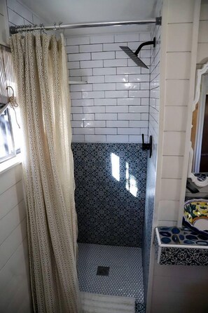 Bagno con pareti piastrellate in bianco, zona doccia, piastrelle decorate sul pavimento e finestra con tenda.