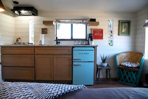 A compact kitchen with a blue Galanz refrigerator, wooden cabinets, a small island with a window, and a wicker chair.