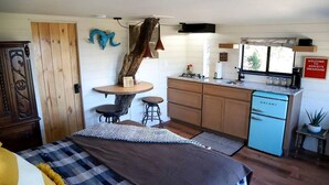 A compact room with a bed, a small dining table with stools, a kitchenette with a blue refrigerator, and a rustic decor including a tree trunk and a ram skull.