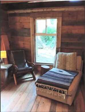 Ein Zimmer in einer Blockhütte mit einem Bett, einem Sessel, einer Lampe und einem Fenster.