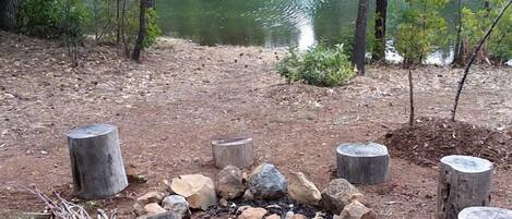 Una fogata rodeada de rocas y tocones de madera, con un lago y árboles al fondo.