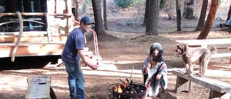 Un entorno rústico al aire libre con un brasero, construcciones de madera y un perro.