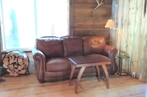 Ein braunes Ledersofa, ein kleiner Holztisch und eine Lampe in einem Raum mit Holzwänden und einem Fenster.