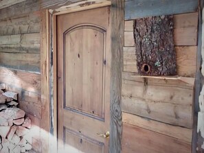 A wooden door with a brass handle, a stack of firewood, and a birdhouse mounted on the wall.