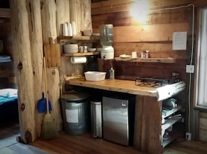 Maalaishenkinen keittiö, jossa on puuseinät, puinen työtaso, pesuallas, jääkaappi ja luuta.