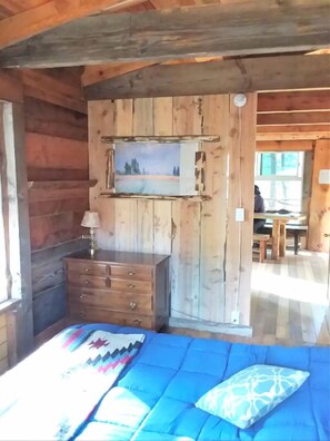 A wooden cabin interior with a bed, dresser, and a painting on the wall.