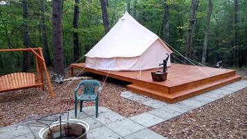 Una carpa con una terraza de madera, un brasero, una silla y un conjunto de columpios en un área boscosa.