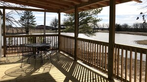 A covered outdoor patio with a table and chairs, wooden railings, and a view of a body of water and trees.