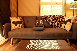 Un sofá marrón con estampado de leopardo y cojines con motivos de animales, un otomán estampado con cebra, y dos copas de vino sobre una mesita.