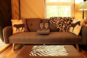 A brown sofa with leopard print and animal-themed cushions, a zebra-patterned ottoman, and two wine glasses on a side table.