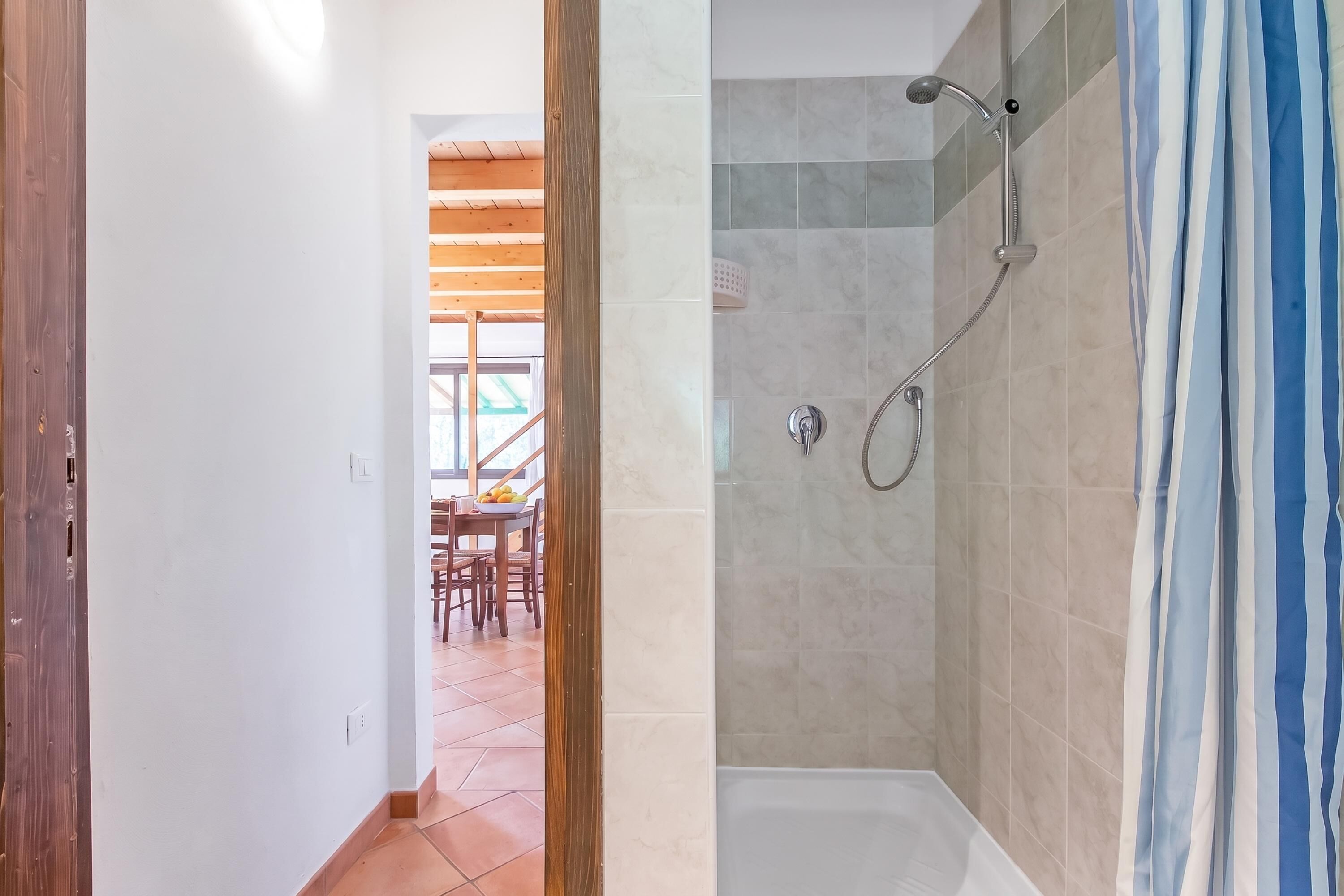 A bathroom with a shower, a toilet, and a sink. There is a wooden door leading to a dining area with a table and chairs.