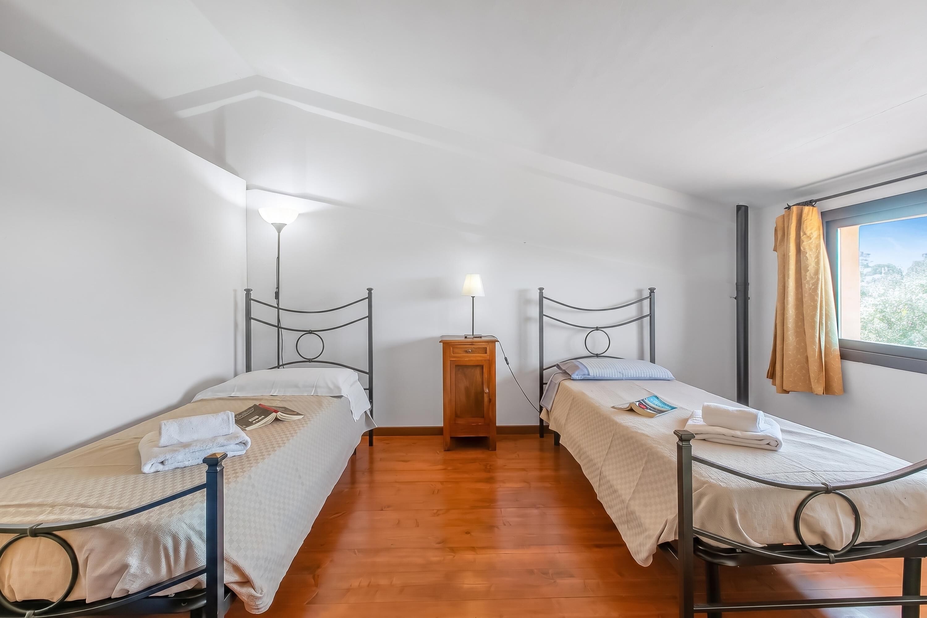 A room with two single beds, a nightstand, a lamp, and a window with curtains.
