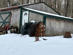 A wooden shed with a white door, surrounded by snow, with a stack of firewood and a small red mailbox in front.
