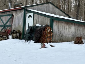 Un abri en bois avec une porte blanche, entouré de neige, devant lequel se trouve une pile de bois de chauffage et une petite boîte aux lettres rouge.
