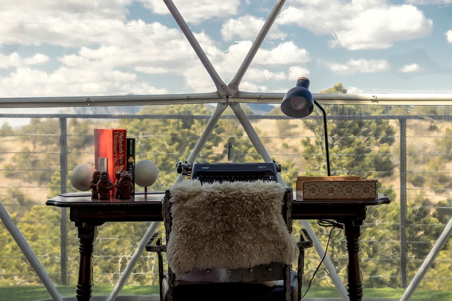 A room with a typewriter, a lamp, and books on a desk by a window with a view of trees.
