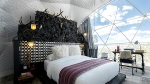 Ein Schlafzimmer mit kariertem Kopfteil, einem großen Bett mit gestreifter Decke, einem Schreibtisch mit Lampe und Blick durch ein großes Fenster.