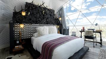 A bedroom with a checkered headboard, a large bed with a striped blanket, a desk with a lamp, and a view through a large window.