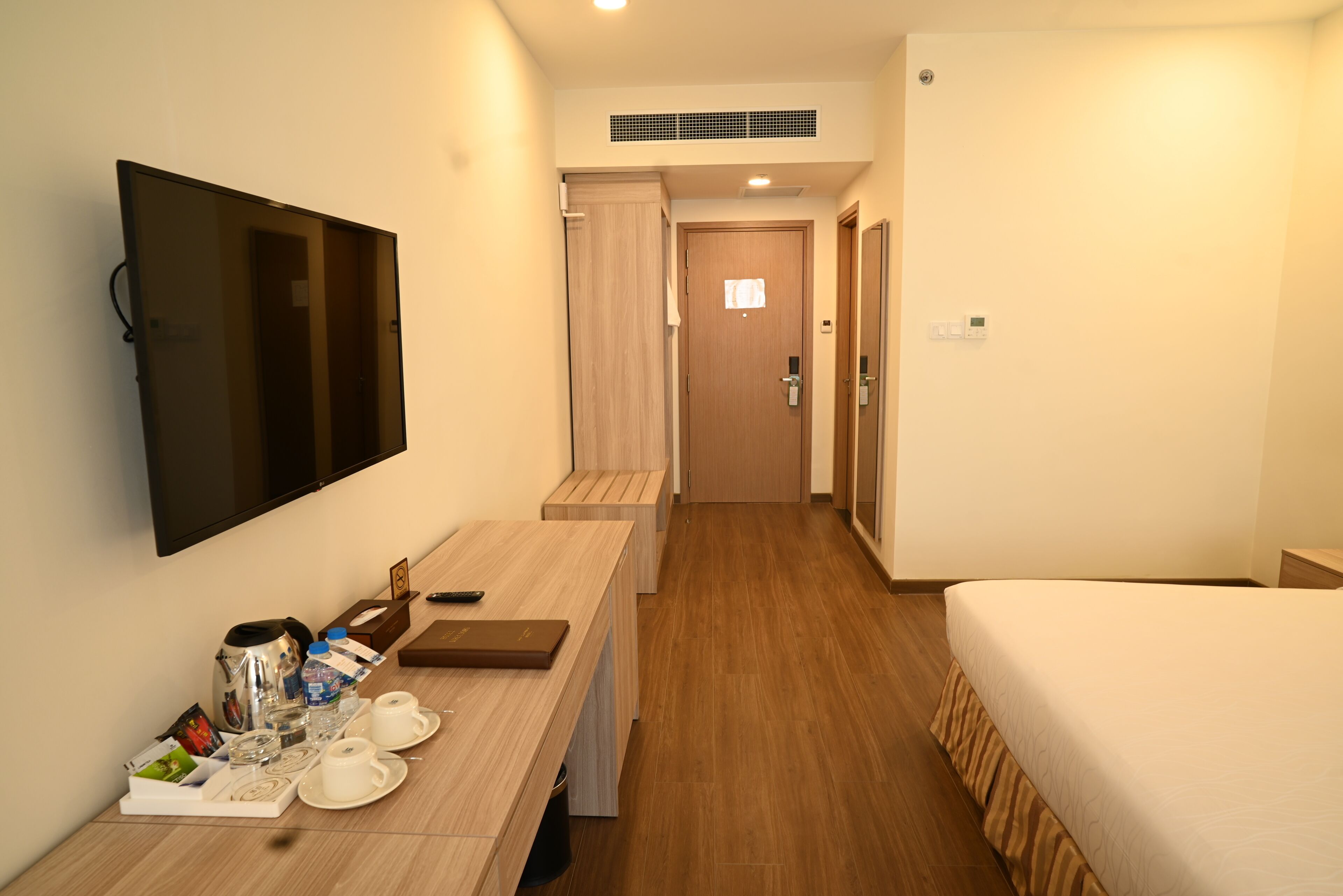 A hotel room with a bed, a TV mounted on the wall, a desk with a coffee maker, cups, and a tray, and a wooden floor.