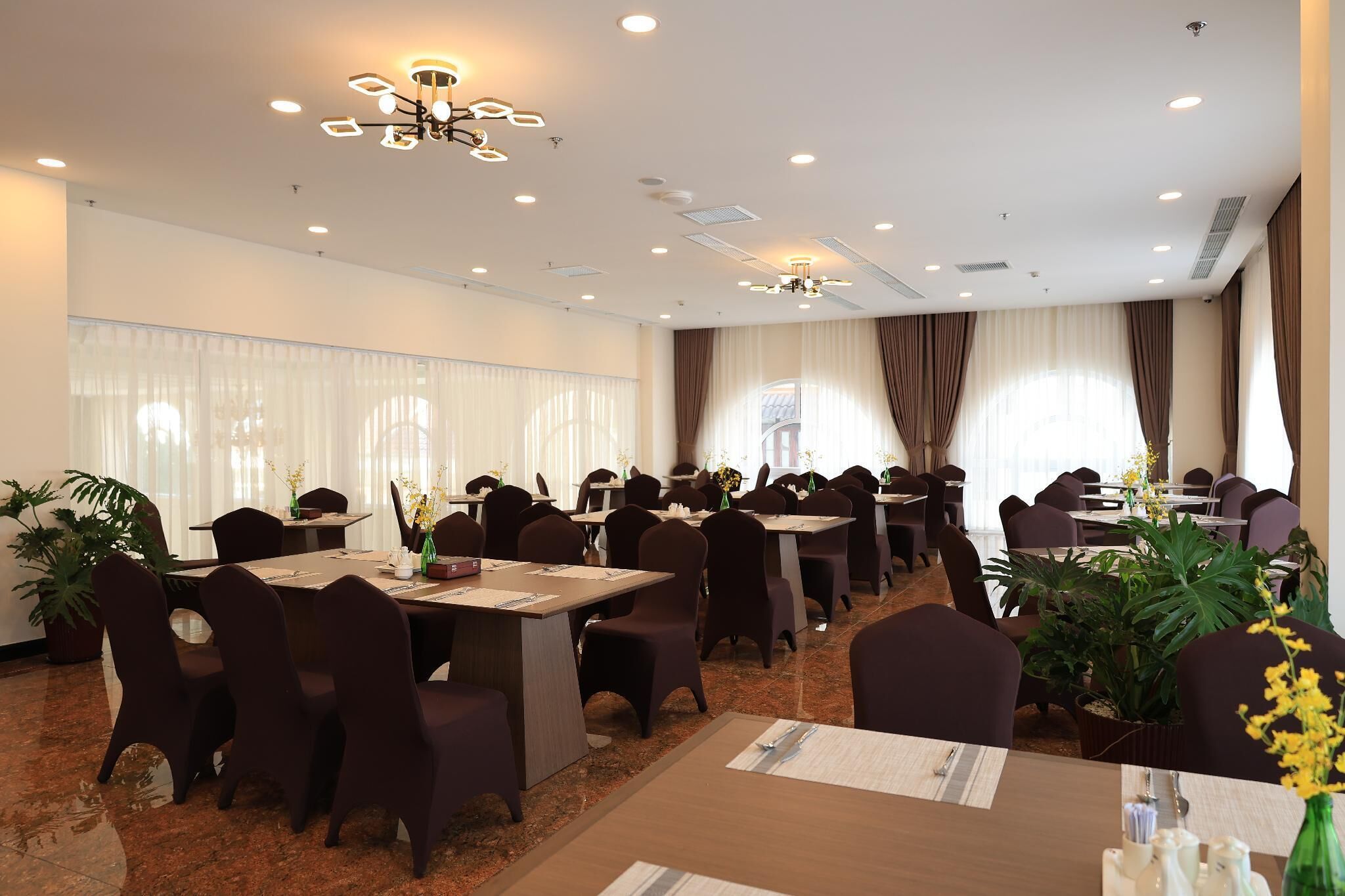 A conference room with multiple tables and chairs, ceiling lights, and large windows with curtains.