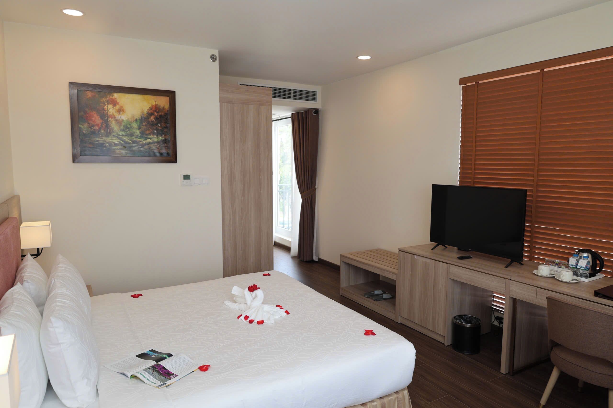 Une chambre d’hôtel avec un lit, un bureau avec une télévision, un tableau au mur et une fenêtre avec des stores.