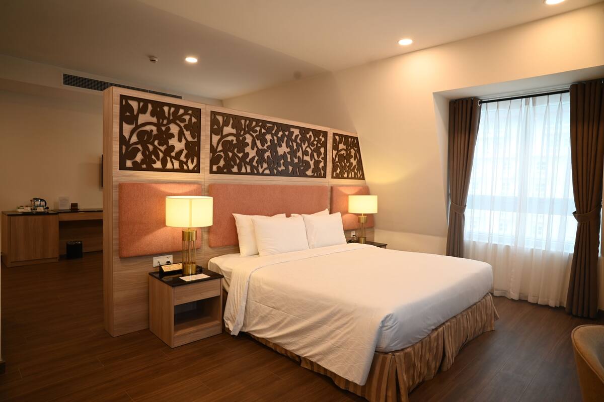 Une chambre d’hôtel avec un grand lit, deux lampes de chevet, une tête de lit en bois, un bureau et une fenêtre avec des rideaux.