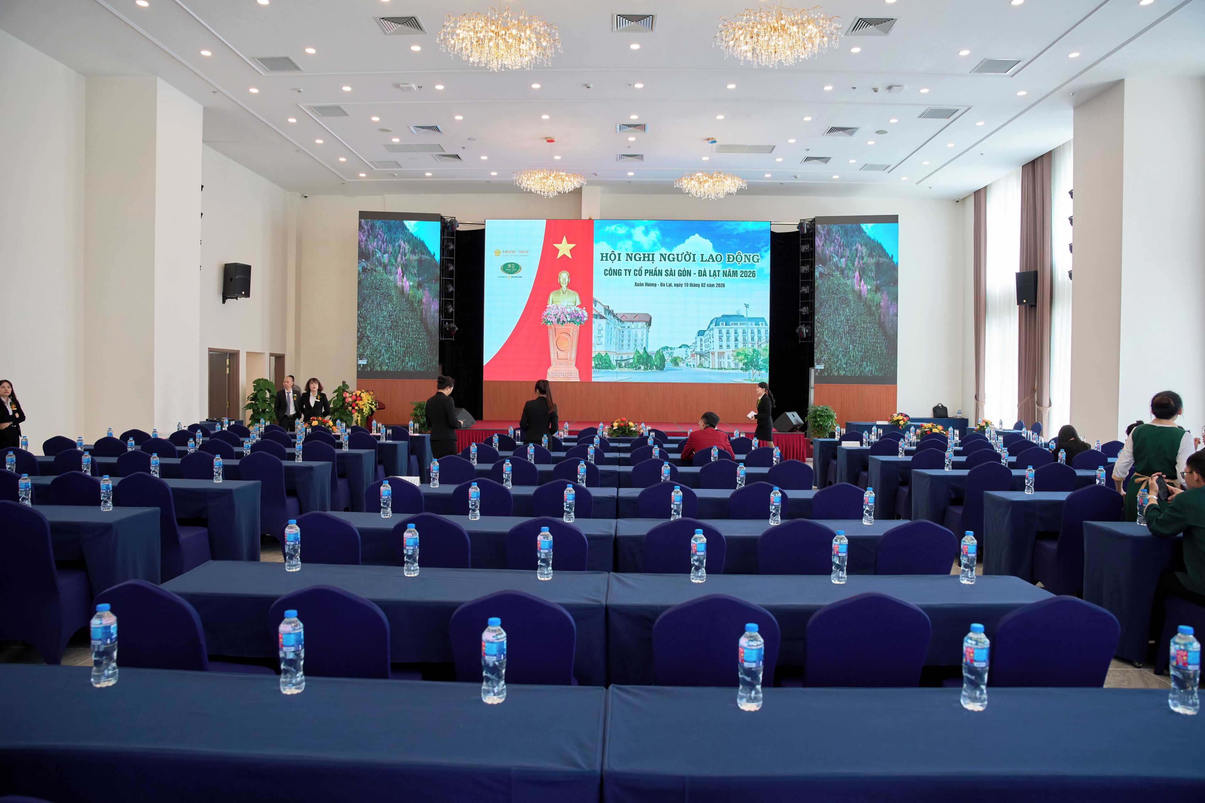 Une salle de conférence avec des rangées de tables, des bouteilles d’eau et un grand écran sur lequel est projetée une présentation.