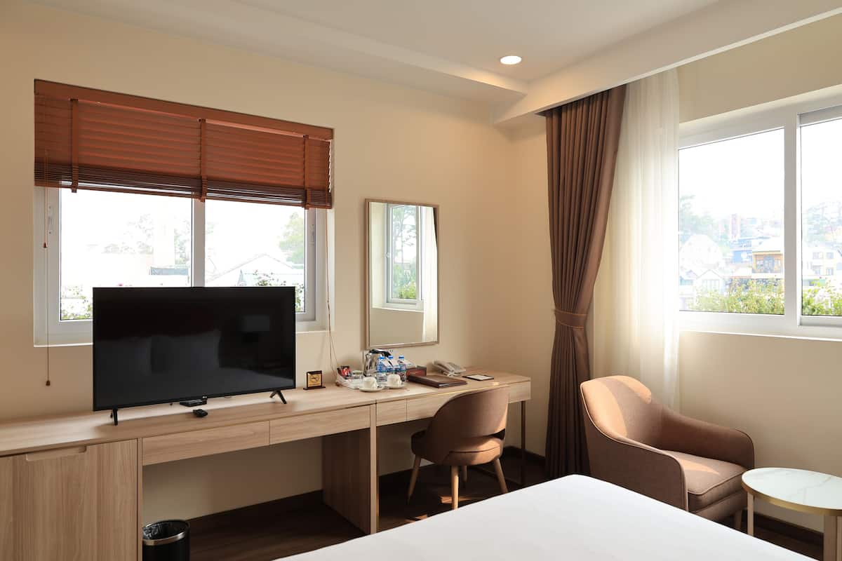 Une chambre d’hôtel avec un lit, un bureau avec une télévision, une chaise, une petite table et des fenêtres avec des rideaux.