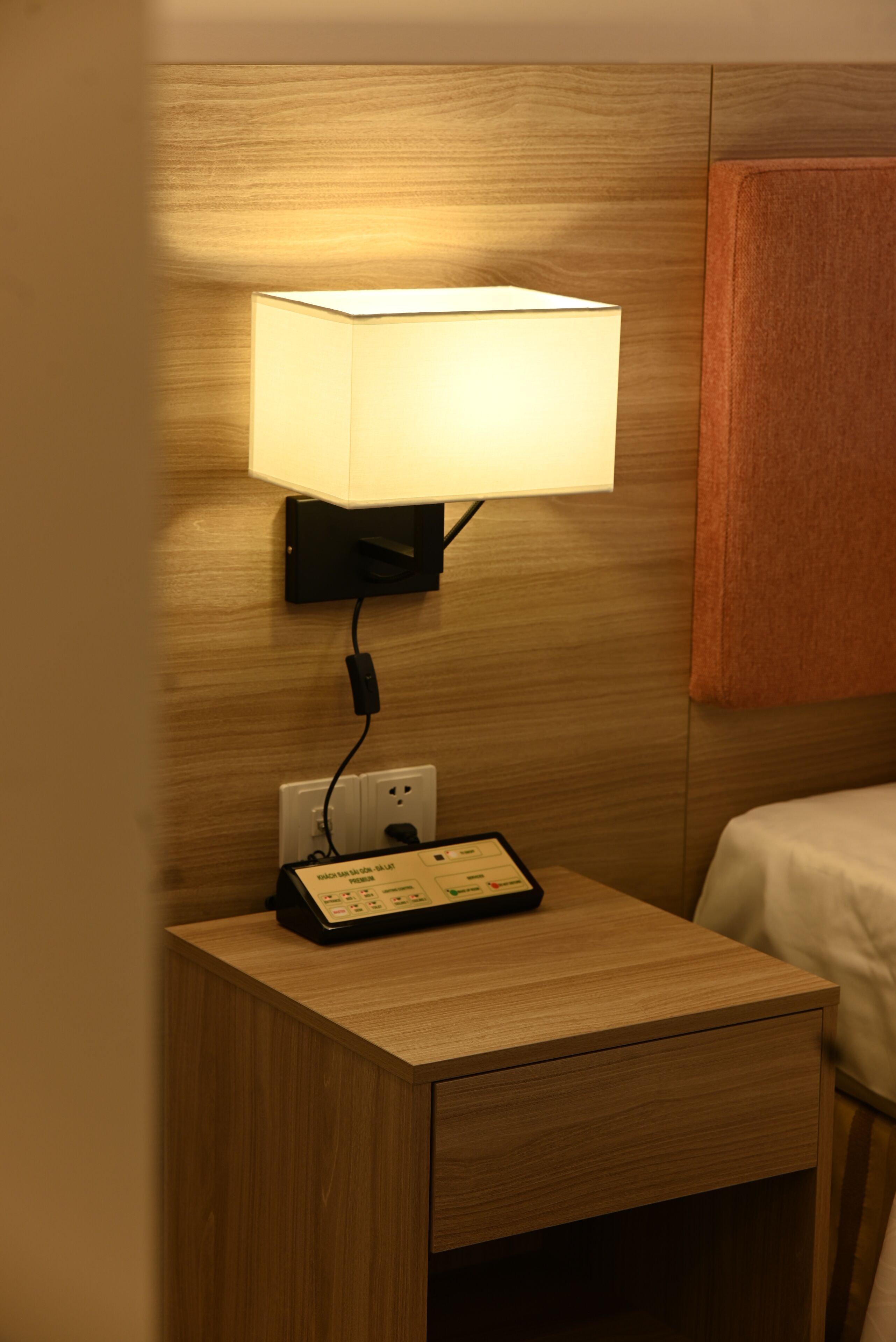 A wooden bedside table with a lamp, a power strip, and a control panel.