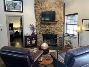 A living room with a stone fireplace, leather sofas, a coffee table, a TV mounted on the wall, and a bookshelf. - Wimberley Romance: Escape to a Cozy Cabin Nestled in Nature, TX (Wimberley)