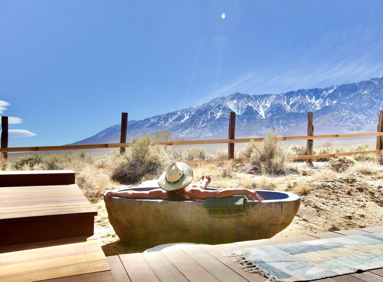 A wooden deck with a concrete bathtub and a hat on it, overlooking a desert landscape with mountains in the background.