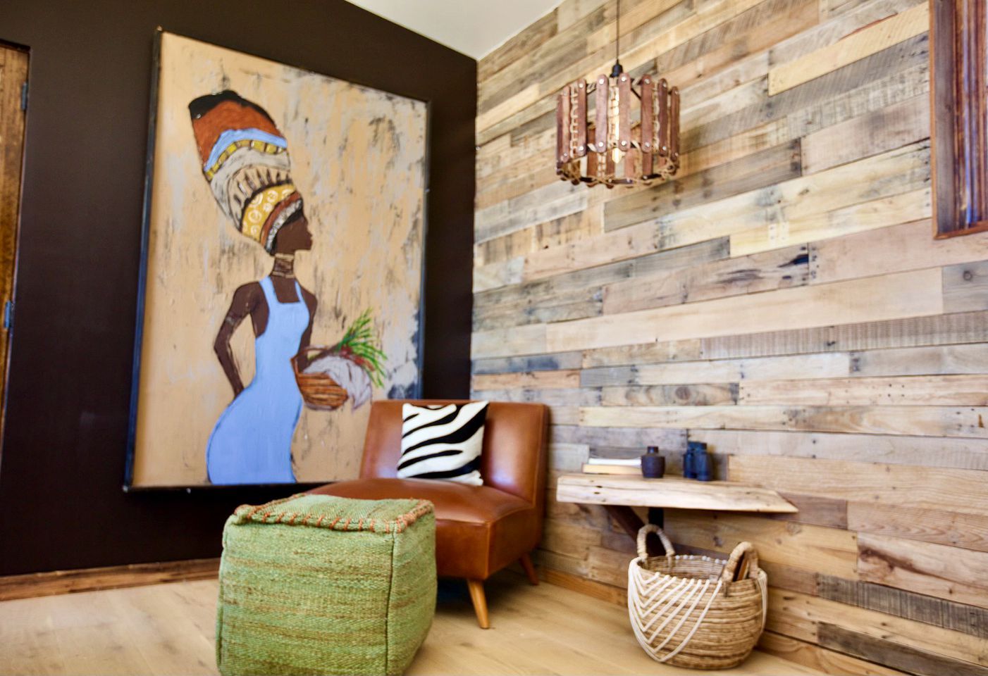 A room with a wooden wall, a large painting, a brown sofa, a zebra-patterned pillow, a woven ottoman, a woven basket, and a hanging light fixture.