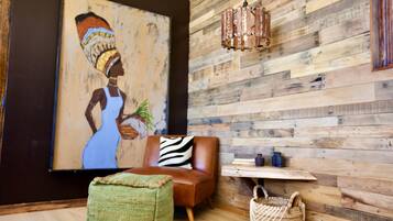 A room with a wooden wall, a large painting, a brown sofa, a zebra-patterned pillow, a woven ottoman, a woven basket, and a hanging light fixture.