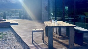 A wooden deck with tables and benches, a glass wall reflecting mountains, and a modern building with a sloped roof.