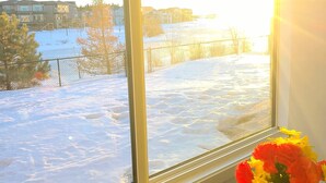A window view of a snowy landscape with houses in the background, a table with a vase of flowers, and a bottle. - Swithome Properties Cozy Suite With Pond View, Near YEG Airport (Edmonton)