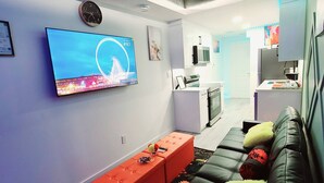 A modern living room with a flat-screen TV, a sofa, a red ottoman, and a kitchen area in the background. - Swithome Properties Cozy Suite With Pond View, Near YEG Airport (Edmonton)