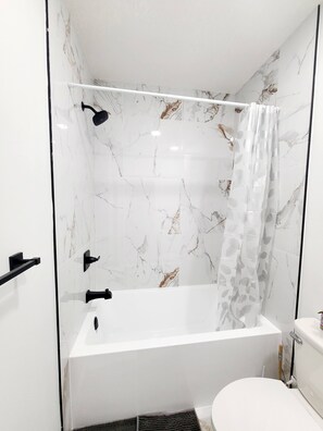 A bathroom with a white bathtub, a toilet, and a shower curtain with a leaf pattern. - Swithome Properties Cozy Suite With Pond View, Near YEG Airport (Edmonton)