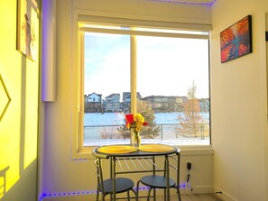 A room with a large window, a small table with chairs, a vase with flowers, and a painting on the wall. - Swithome Properties Cozy Suite With Pond View, Near YEG Airport (Edmonton)