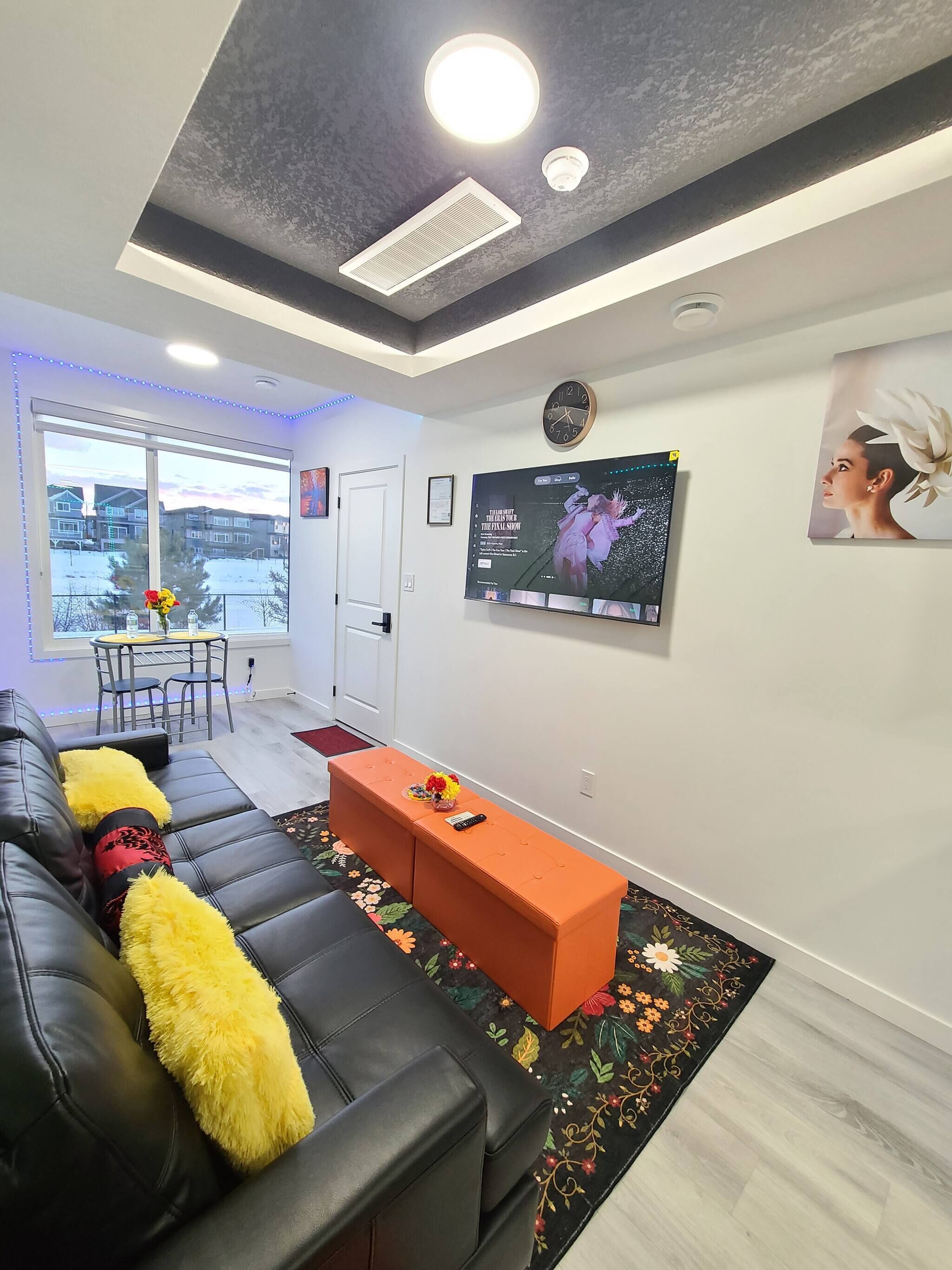 A modern living room with a black leather sofa, an orange ottoman, a flat-screen TV, and a large window with a view of buildings.