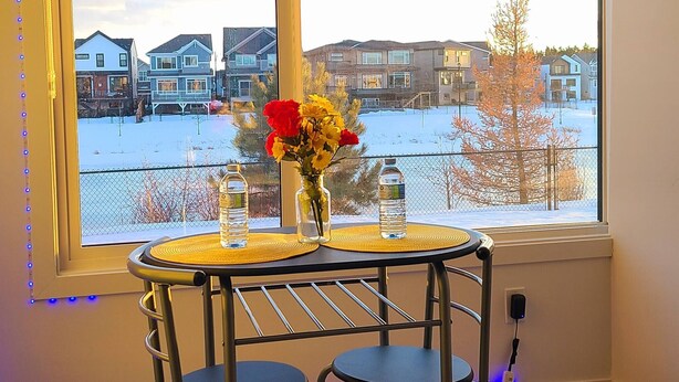 A room with a large window, a table with two chairs, a vase with flowers, and a bottle of water. - Swithome Properties Cozy Suite With Pond View, Near YEG Airport (Edmonton)