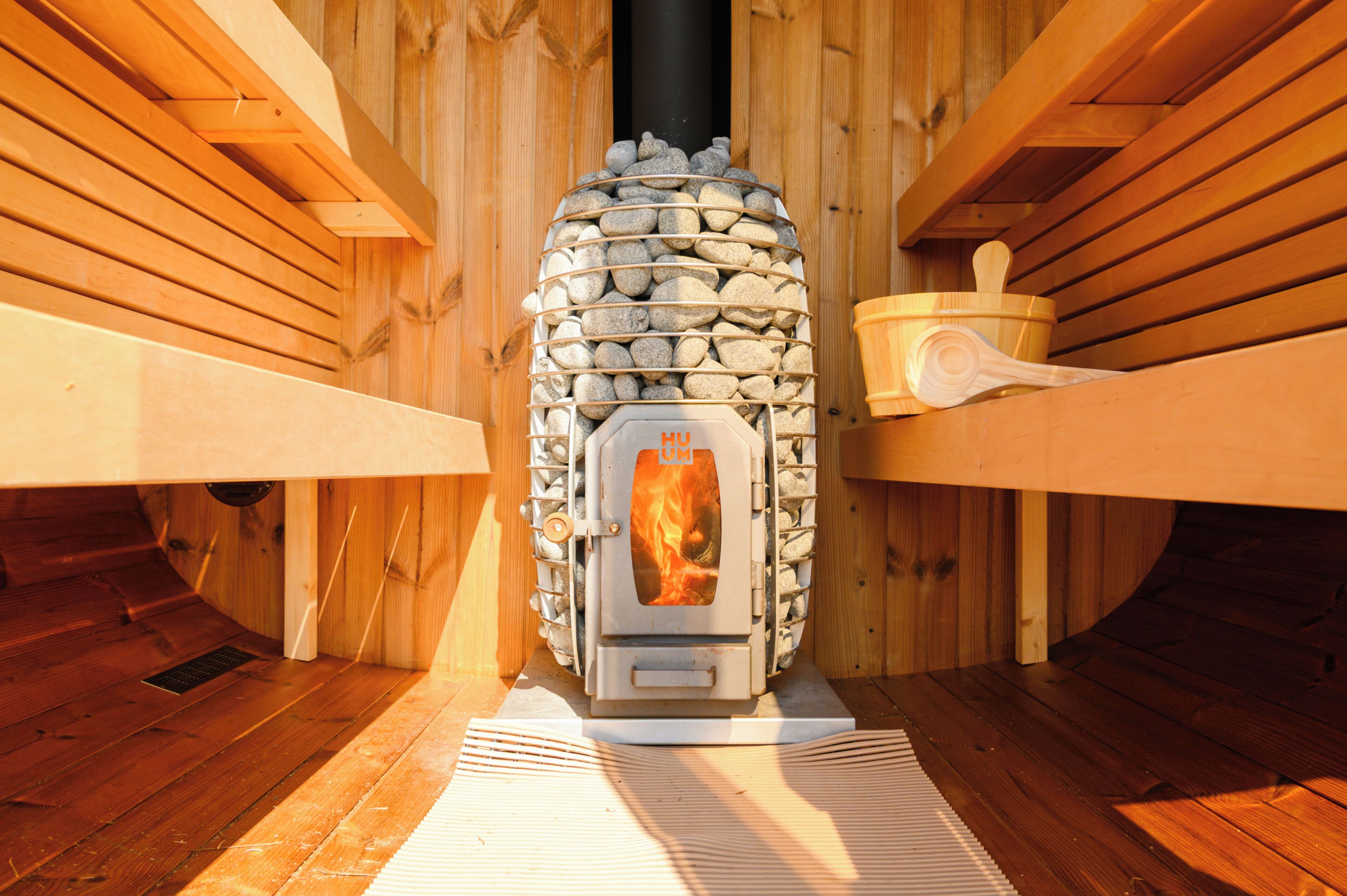 Una estufa de leña encendida, ubicada en una habitación de madera con paredes de listones y suelo de madera.