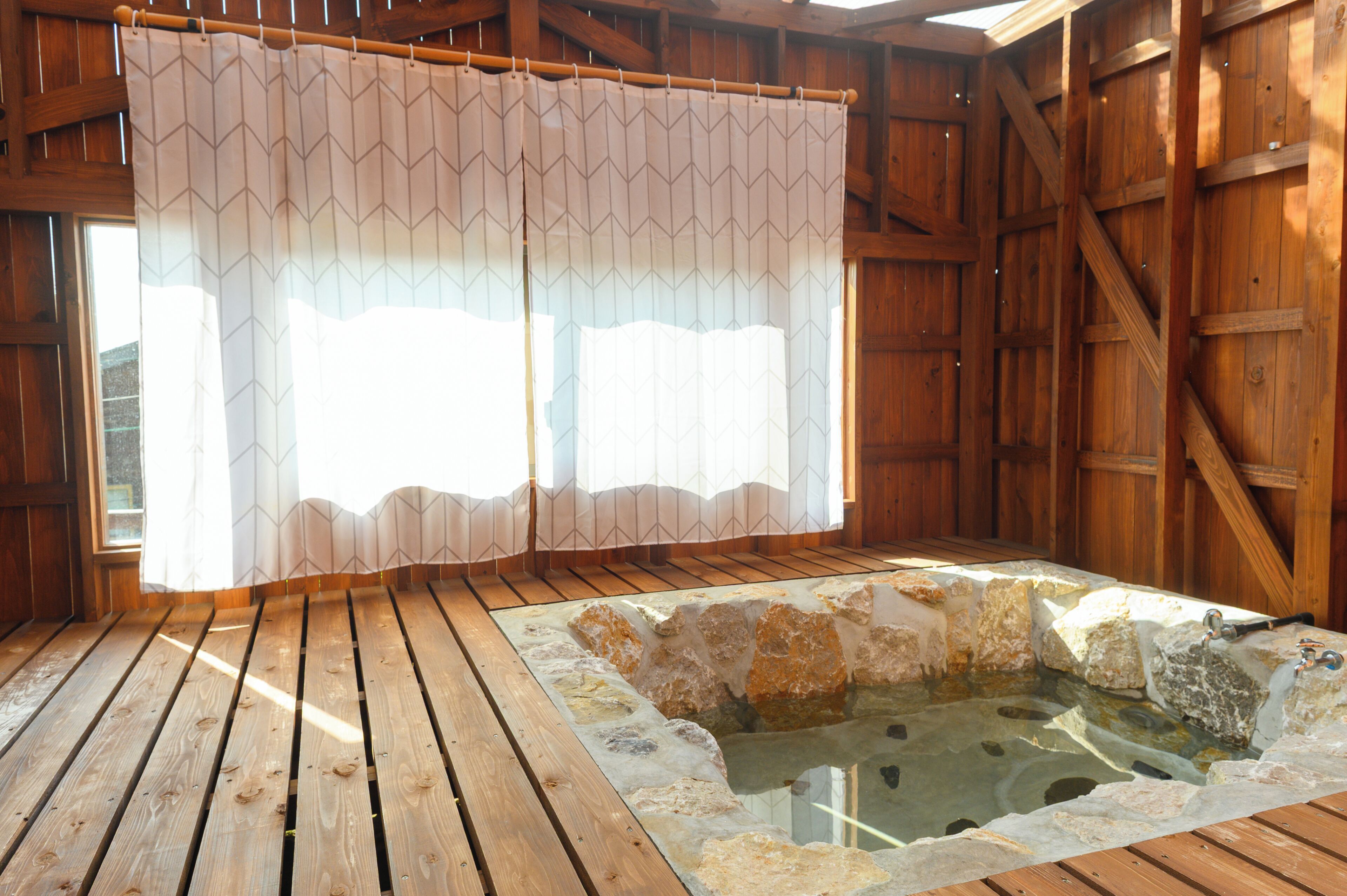 A wooden room with a hot spring, wooden flooring, and a window with a curtain.