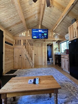 Ein Holzinterieur mit einem an der Decke befestigten Fernseher, einem Esstisch mit einer Fernbedienung und einer Küchenzeile.