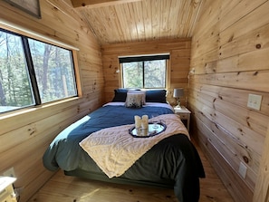 A wooden cabin bedroom with a bed, bedside table, and a window. - Incredible Modern Tiny Log Cabin near the Ammonoosuc River for a Romantic Getaway in New Hampshire (Bath)